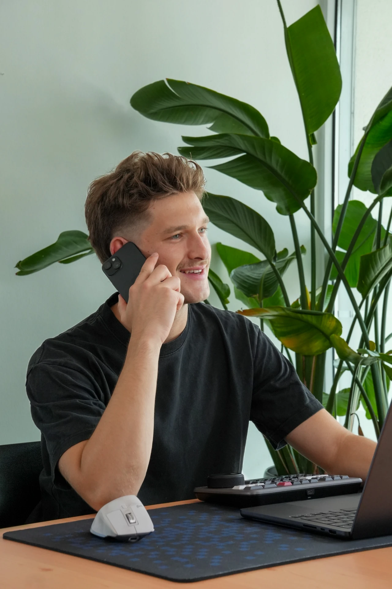 Noah Wierer telefoniert an seinem Arbeitsplatz mit einer großen Monstera-Pflanze im Hintergrund